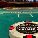 A custom-designed casino poker chip for the 'Skyline Dealer Poker Room' resting on a green felt blackjack table, with a blurred casino interior in the background.